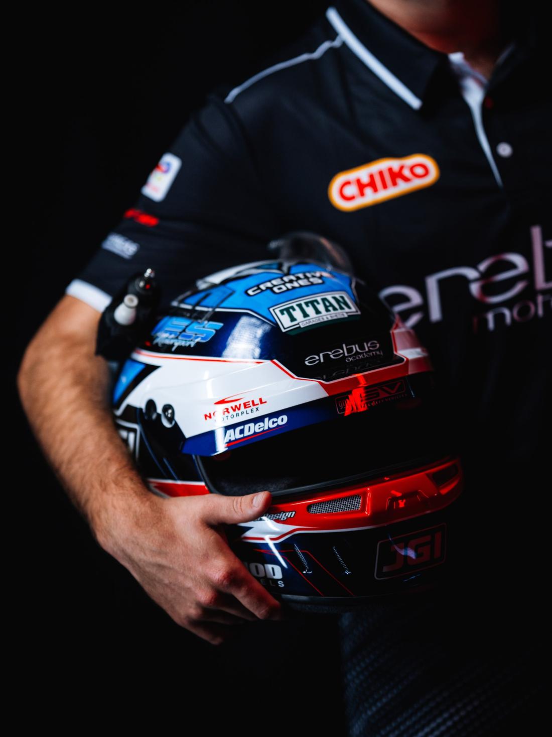 A driver in an Erebus Motorsport polo holds his race helmet under his arm, face out of frame